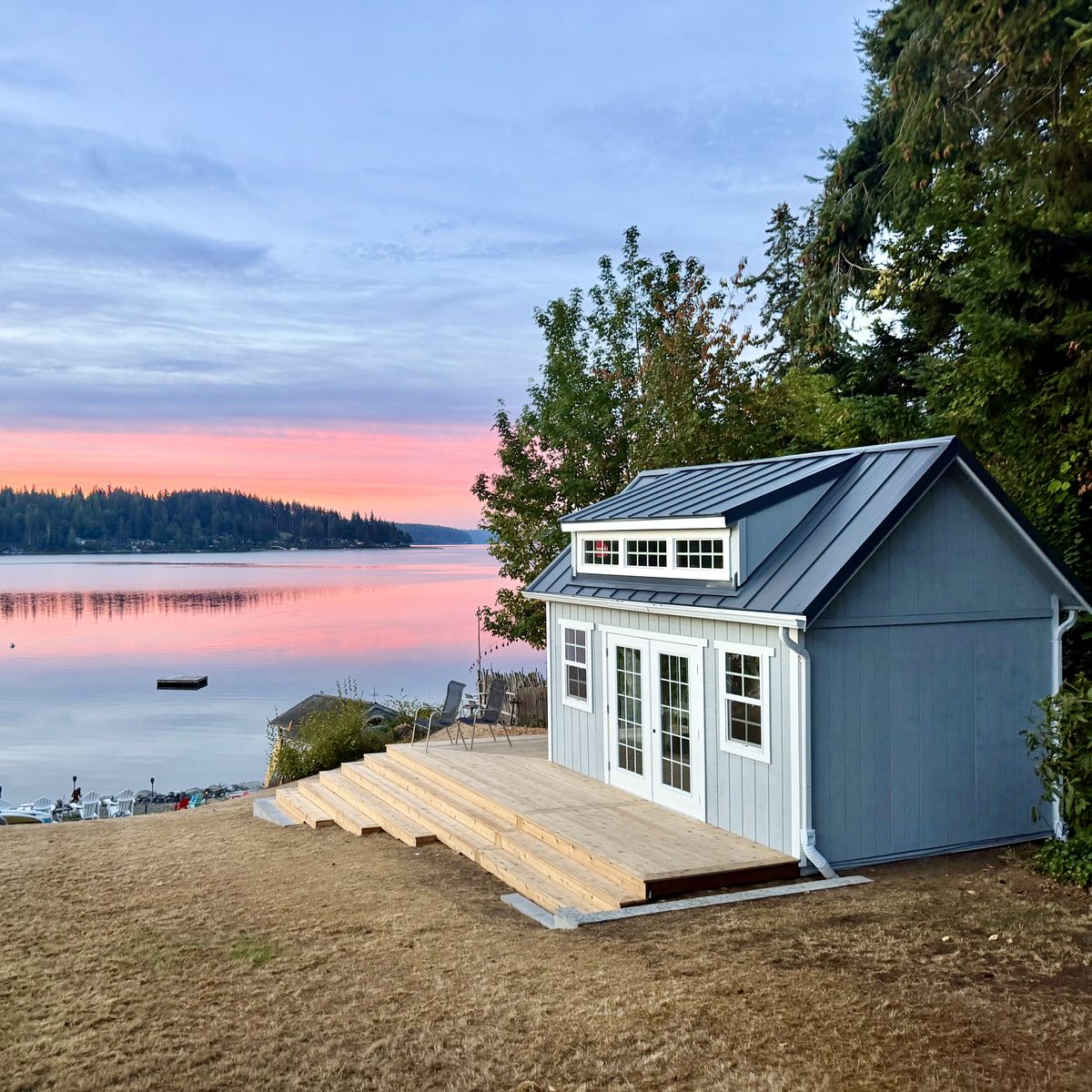 Grey backyard studio with a metal roof and French doors, opening onto a new wood deck above the water at sunset